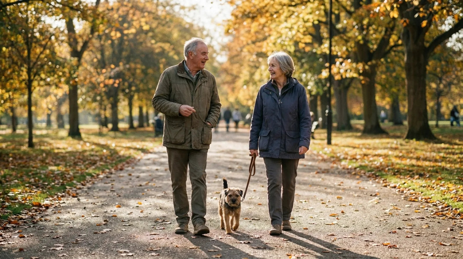 An older couple walking a small dog along a park path in autumn light, talking to each other rather than watching the ground