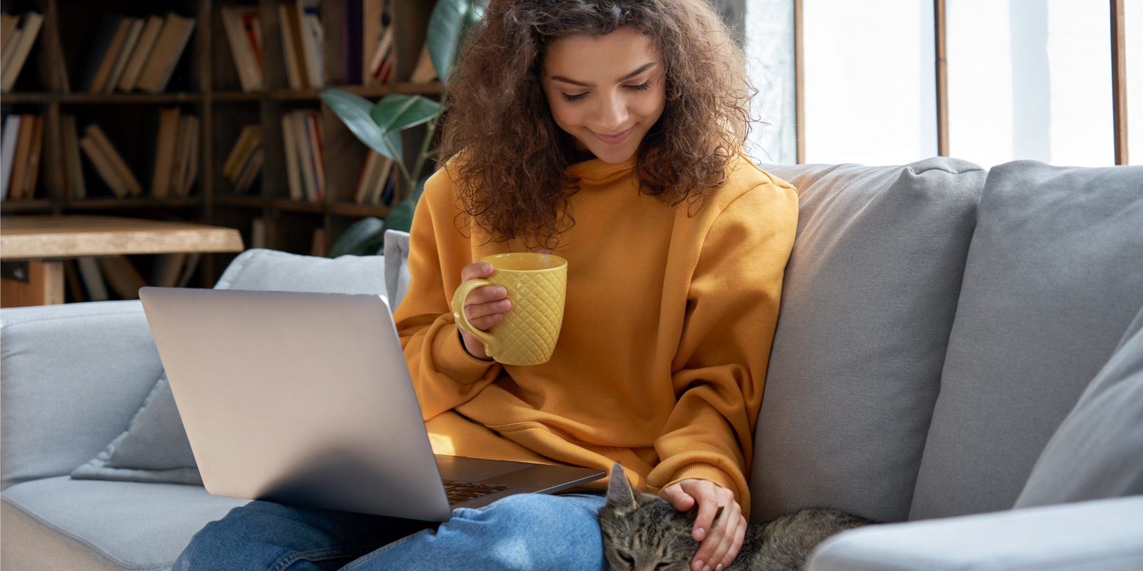 Happy hispanic teen girl relaxing on couch holding laptop playing with cat.