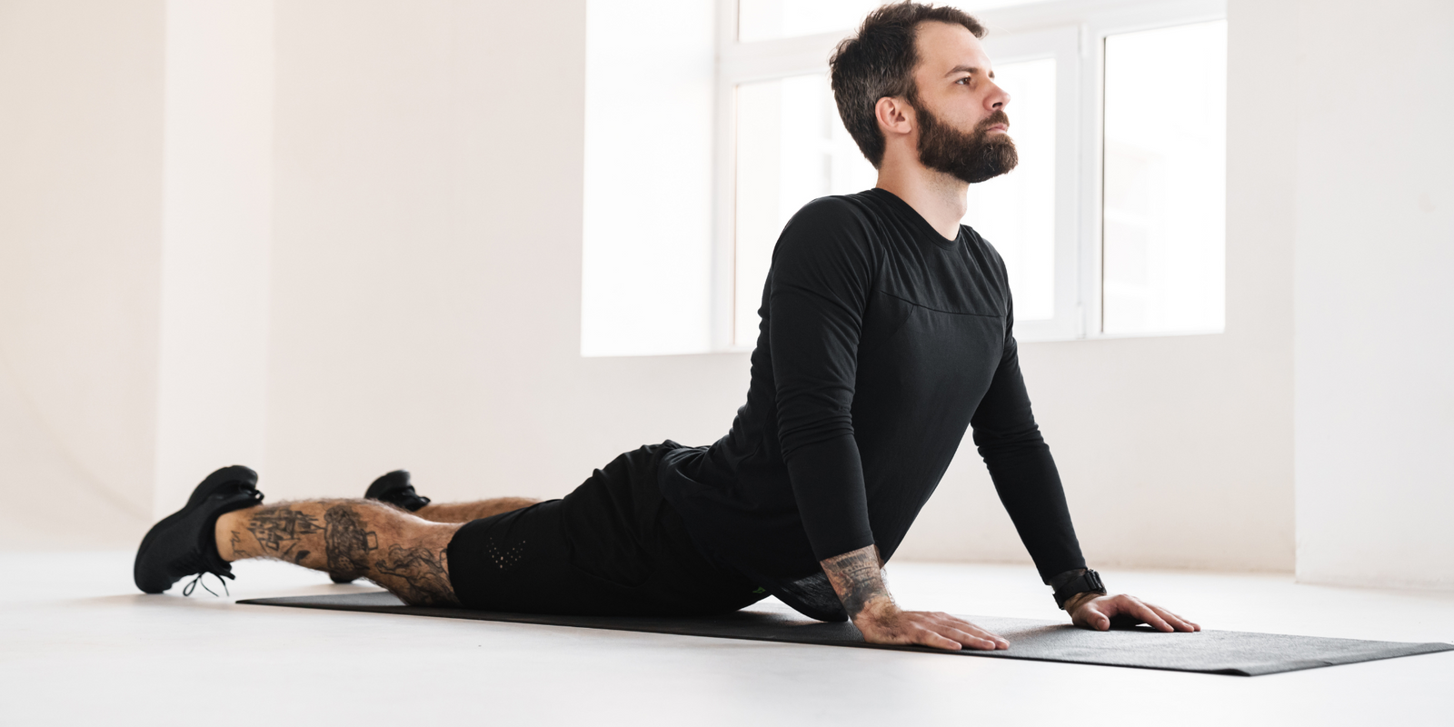 Young brunette bearded man in sportswear stretching while laying on a fitness mat indoors