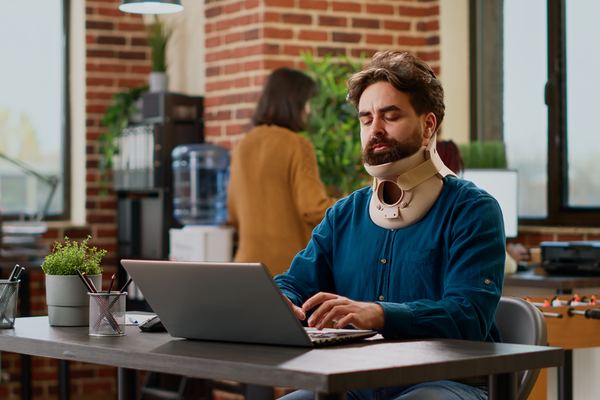 Hurt businessman wearing cervical neck brace at office job, working on project with pain and injury. Person with physical tension and collar foam trying to cure after accident, suffering at work.