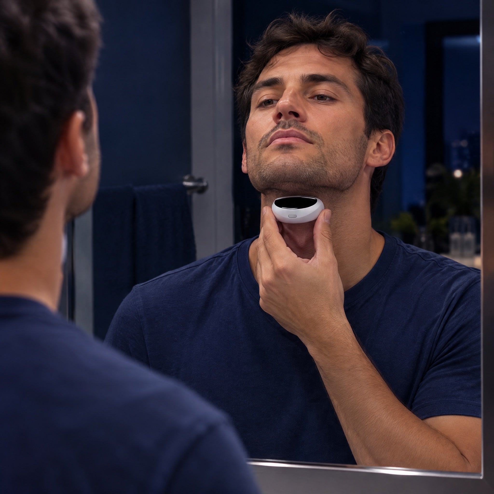A man in a bathroom mirror holding a small silver pod just under his jaw.