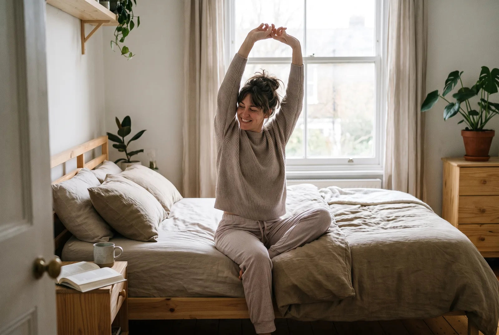A woman sitting on the edge of her bed in morning light, stretching, rested